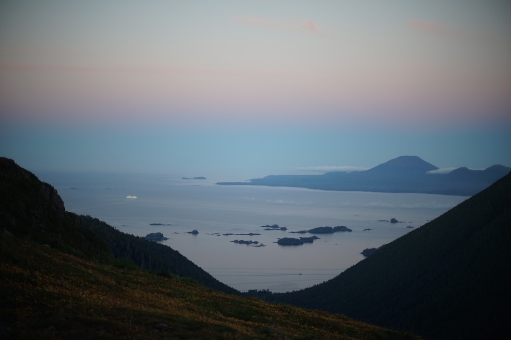 the sky is pink, purple, blue, pale yellow, silver. a conical volcanic mountain stretches its broad base into a pale silver and blue sea. soft green slopes in the early morning dusk frame the ocean, the conical volcanic peak. a white boat, a massive cruise ship, looks tiny in the midst of all this earth.