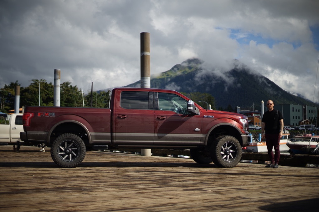 a large 4 door pickup truck sits on a wooden dock. behind it, a mountain is mostly hidden by cloud. 